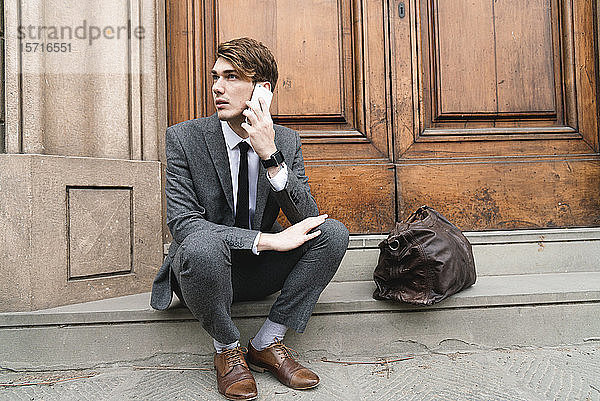 Porträt eines jungen Geschäftsmannes am Telefon  der auf den Stufen des Hauseingangs sitzt