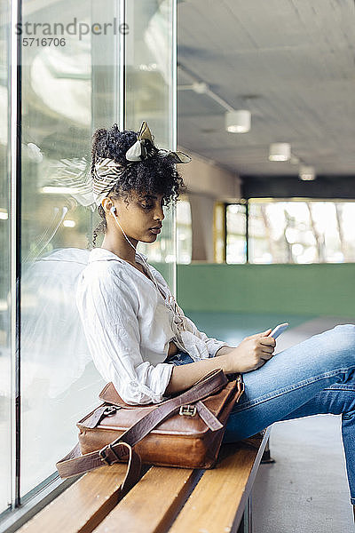 Porträt einer jungen Frau mit Ohrstöpseln  die auf einer Bank sitzt und auf ein Mobiltelefon schaut