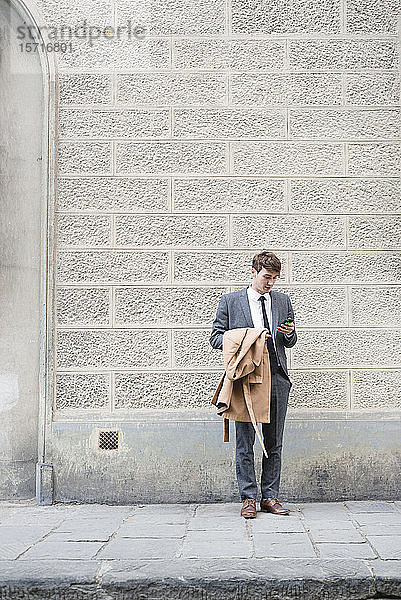 Junger Geschäftsmann steht auf Bürgersteig und schaut auf Smartphone