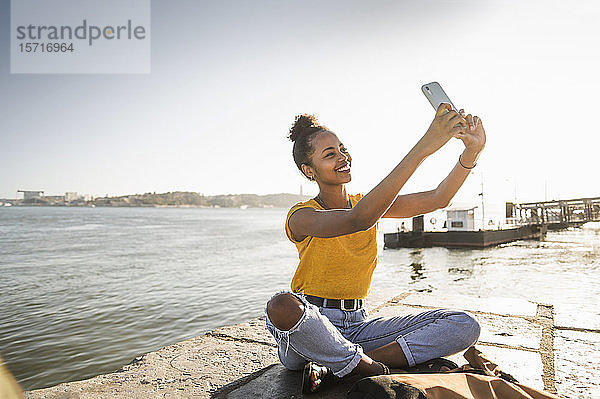 Glückliche junge Frau sitzt auf dem Pier am Wasser und macht einen Selfie  Lissabon  Portugal