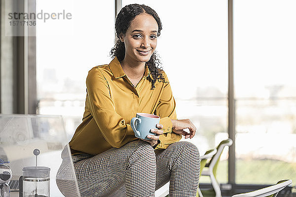 Porträt einer lächelnden jungen Geschäftsfrau  die im Büro auf dem Schreibtisch sitzt und eine Kaffeepause macht