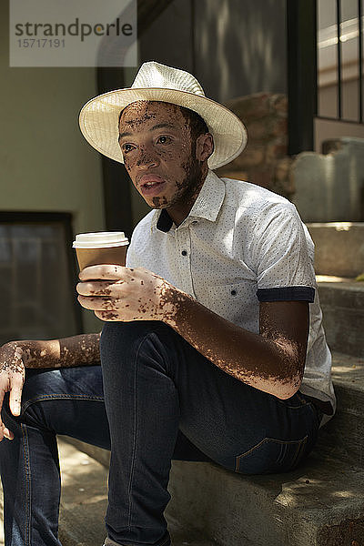 Porträt eines jungen Mannes mit Vitiligo  der einen Hut trägt und einen Kaffee zum Mitnehmen auf der Treppe sitzt