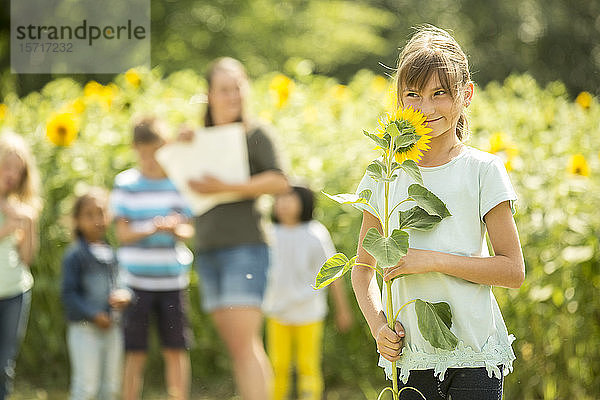 Kleines Mädchen hält Sonnenblume  im Hintergrund stehen Lehrer und Freunde