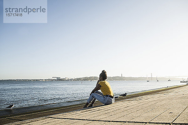 Rückansicht einer jungen Frau  die bei Sonnenuntergang am Wasser sitzt  Lissabon  Portugal