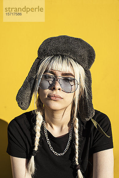 Porträt einer jungen Frau mit gefärbten Haaren  die eine Mütze und eine Sonnenbrille gegen eine gelbe Wand trägt