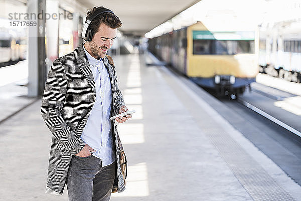 Junger Geschäftsmann mit Handy und Kopfhörern am Bahnhof