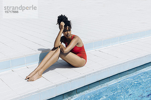 Porträt einer nachdenklichen jungen Frau in rotem Badeanzug am Poolufer sitzend