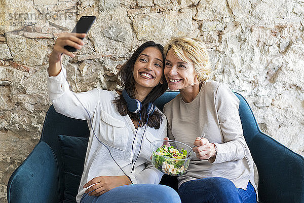 Zwei glückliche Kollegen sitzen zur Mittagszeit auf der Couch und nehmen sich mit dem Smartphone ein
