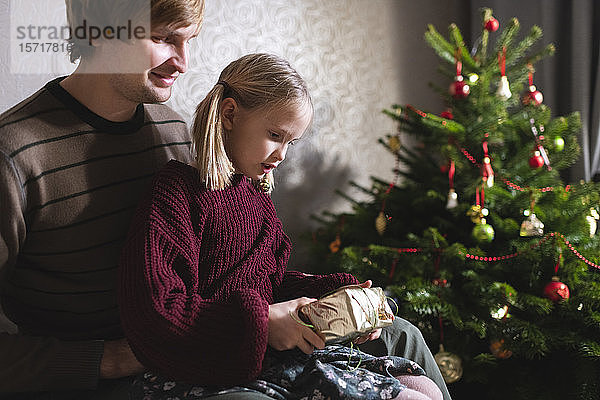Blondes Mädchen sitzt auf dem Schoß des Vaters und öffnet Weihnachtsgeschenk