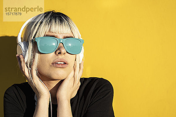 Porträt einer jungen Frau mit blauer Sonnenbrille  die vor einer gelben Wand Musik hört