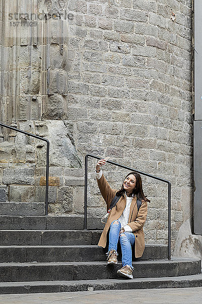 Lächelnde junge Frau  die auf einer Treppe in der Stadt sitzt und ein Selfie nimmt  Barcelona  Spanien