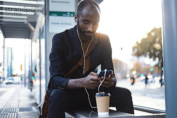 Junger Geschäftsmann sitzt abends mit Kopfhörern und Smartphone an der Straßenbahnhaltestelle