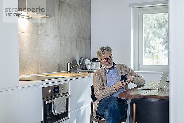 Reifer Mann benutzt Handy und Laptop in der Küche zu Hause