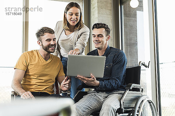 Junger Geschäftsmann im Rollstuhl zeigt Kollegen im Büro seinen Laptop