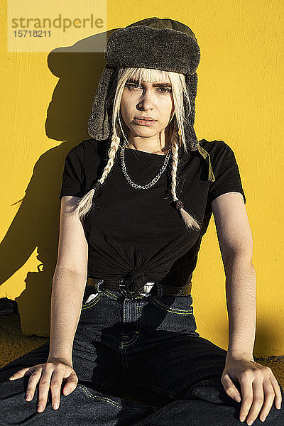 Bildnis einer jungen Frau mit blonden Zöpfen und Mütze vor gelber Wand sitzend