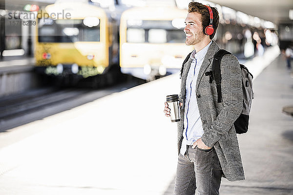Lächelnder junger Geschäftsmann mit Kaffeetasse und Kopfhörer am Bahnhof