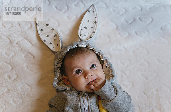 Bildnis eines kleinen Mädchens in einem Kaninchen-Kapuzenpulli mit Finger im Mund auf einer weißen Decke liegend