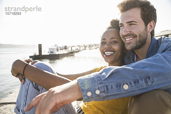 Porträt eines glücklichen jungen Paares  das am Pier am Wasser sitzt  Lissabon  Portugal