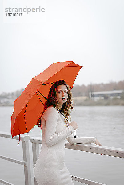 Porträt einer jungen Frau mit rotem Regenschirm  die sich an einem Regentag an ein Geländer lehnt