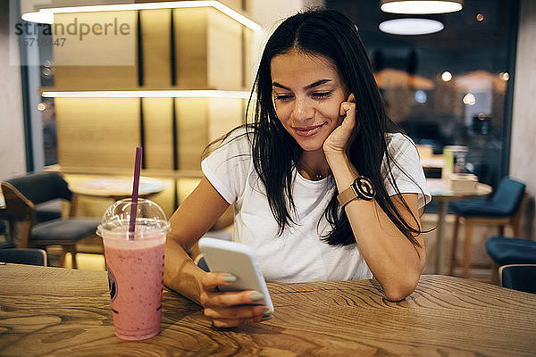 Schwarzhaarige Frau trinkt einen Smoothie und benutzt ein Smartphone im Café
