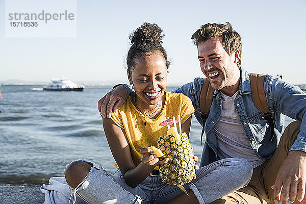 Glückliches junges Paar sitzt am Pier am Wasser mit einer Ananas  Lissabon  Portugal