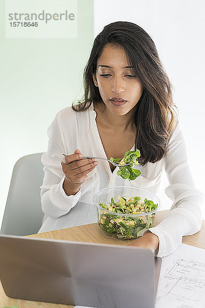 Porträt eines jungen Architekten  der am Schreibtisch gemischten Salat isst und auf seinen Laptop schaut