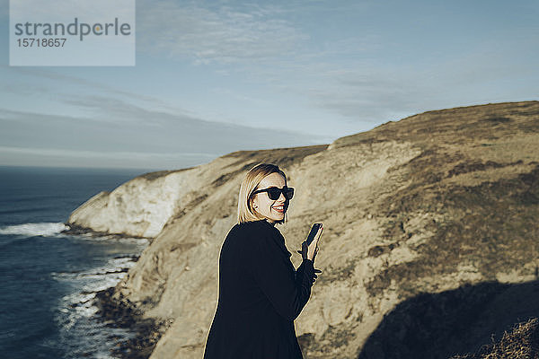 Rückansicht einer jungen blonden Frau mit Smartphone an der Küste