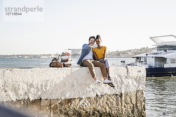 Glückliches junges Paar sitzt auf dem Pier am Wasser und macht einen Egoisten  Lissabon  Portugal