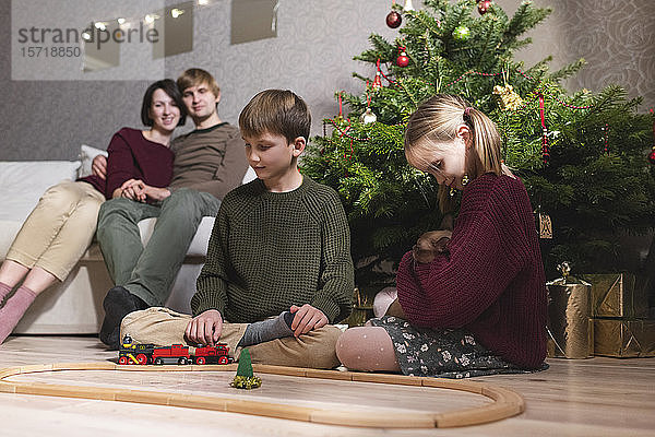 Junge und Mädchen mit Katze  spielen mit der Spielzeugeisenbahn auf einer Holzeisenbahn in der Nähe des Weihnachtsbaums und die Eltern sitzen auf dem Sofa hinter ihren Kindern