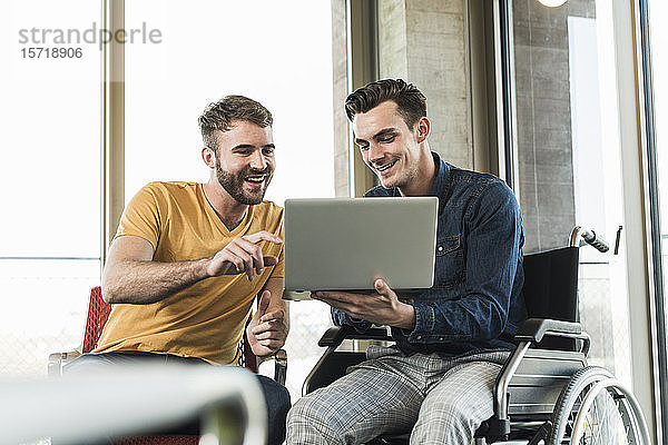 Junger Geschäftsmann im Rollstuhl zeigt Laptop einem Kollegen im Büro