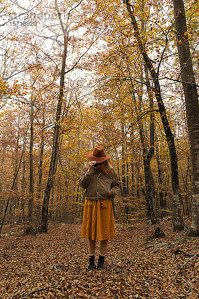 Modische rothaarige junge Frau im herbstlichen Wald  die ihr Gesicht mit einem Hut bedeckt