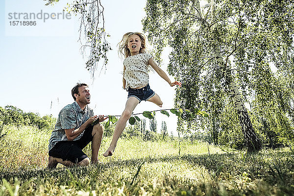Vater mit Tochter springt über Stock in Mohnwiese