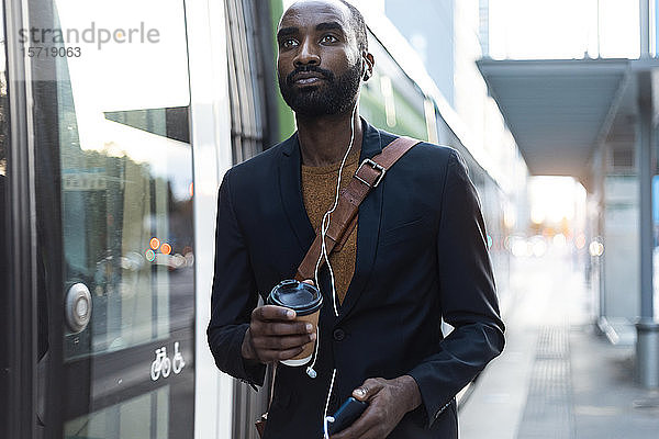 Porträt eines jungen Geschäftsmannes mit Kaffee zum Mitnehmen  Kopfhörer und Smartphone an der Straßenbahnhaltestelle