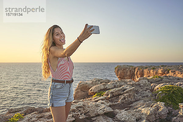 Junge Frau steht auf einer Klippe am Meer  benutzt ein Smartphone  nimmt sich selbst