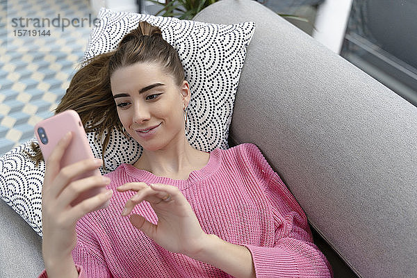 Porträt einer lächelnden jungen Frau  die mit ihrem Handy auf der Couch liegt