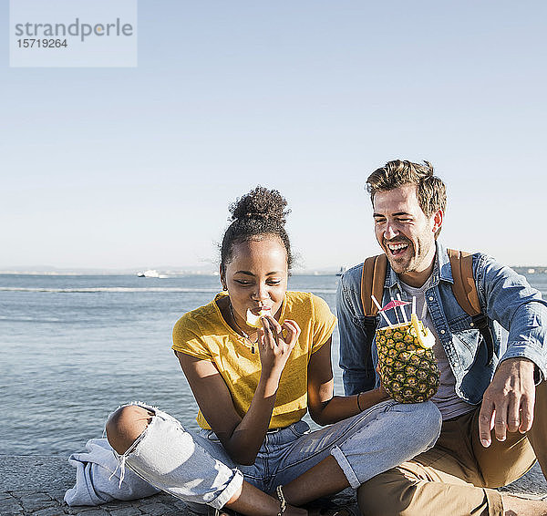 Glückliches junges Paar sitzt am Pier am Wasser mit einer Ananas  Lissabon  Portugal