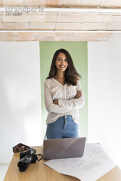 Porträt eines lächelnden jungen Architekten mit Kamera  Laptop und Bauplan auf dem Schreibtisch in einem Atelier