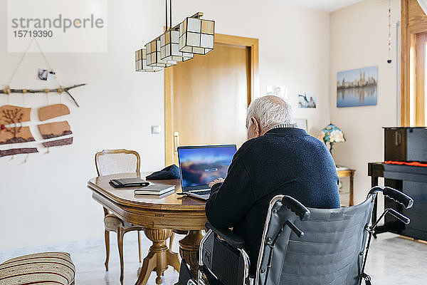 Rückenansicht eines älteren Mannes im Rollstuhl mit Laptop zu Hause
