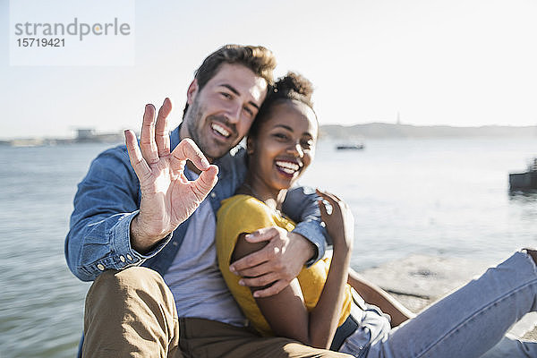 Porträt eines glücklichen jungen Paares  das gestikulierend am Wasser sitzt  Lissabon  Portugal