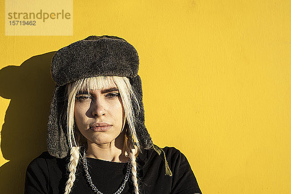 Bildnis einer jungen Frau mit blonden Zöpfen  die eine Mütze gegen eine gelbe Wand trägt