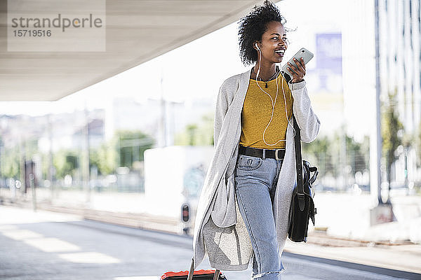 Lächelnde junge Frau unterwegs mit Smartphone am Bahnhof