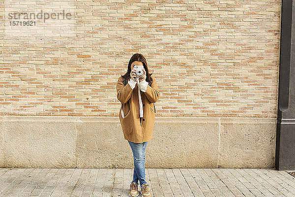Junge Frau mit Kamera beim Fotografieren vor einer Ziegelmauer