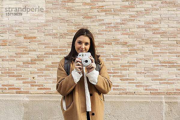 Porträt einer lächelnden jungen Frau mit Kamera vor einer Ziegelmauer