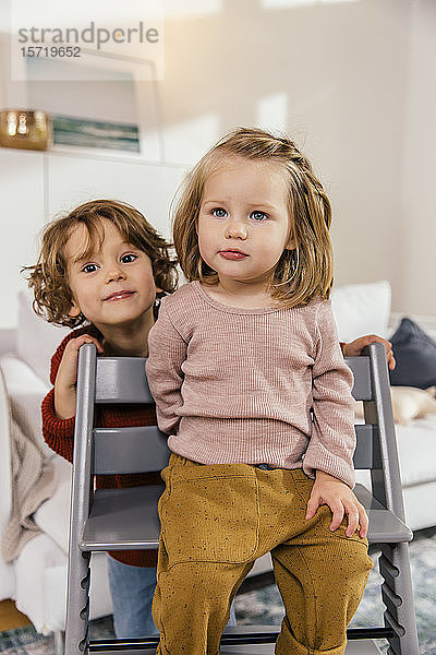 Kleines Mädchen sitzt zu Hause auf Hochstuhl mit Schwester hinter sich