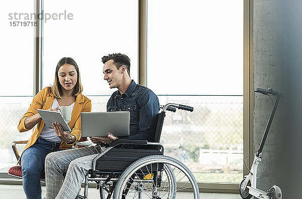 Junger Geschäftsmann mit Laptop im Rollstuhl und Geschäftsfrau mit Tablett im Büro