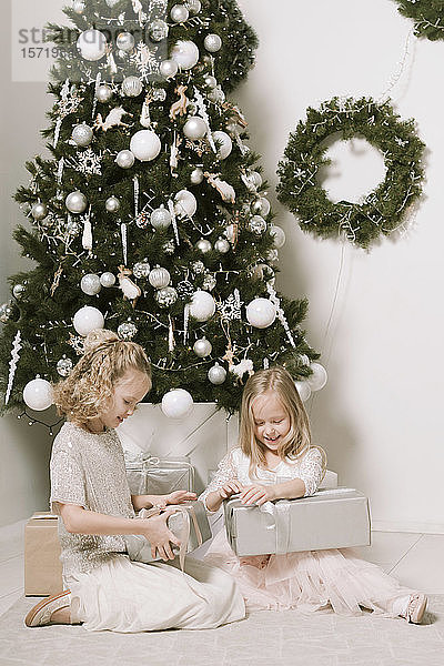 Zwei kleine Mädchen sitzen vor dem Weihnachtsbaum und öffnen Weihnachtsgeschenke