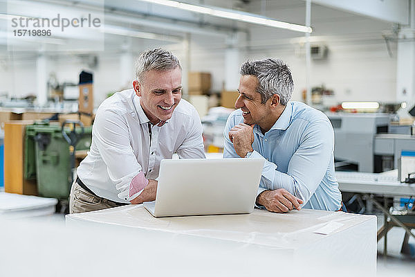 Zwei lächelnde Kollegen am Laptop und diskutieren in einer Fabrik