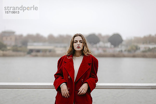 Porträt einer jungen Frau in rotem Mantel  die sich an einem Regentag an die Reling lehnt