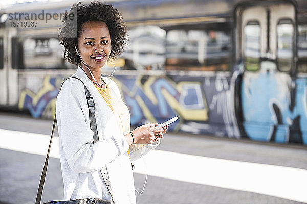 Porträt einer lächelnden jungen Frau mit Handy und Kopfhörer am Bahnhof