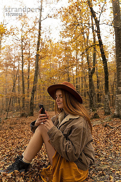Modische rothaarige junge Frau sitzt im herbstlichen Wald und schaut auf ihr Handy
